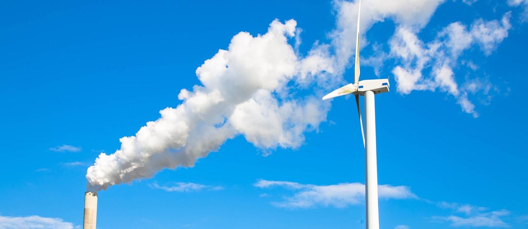 Rauchender Schlot und rotierende Windkraftanlage zueinander gewendet vor blauem Himmel.