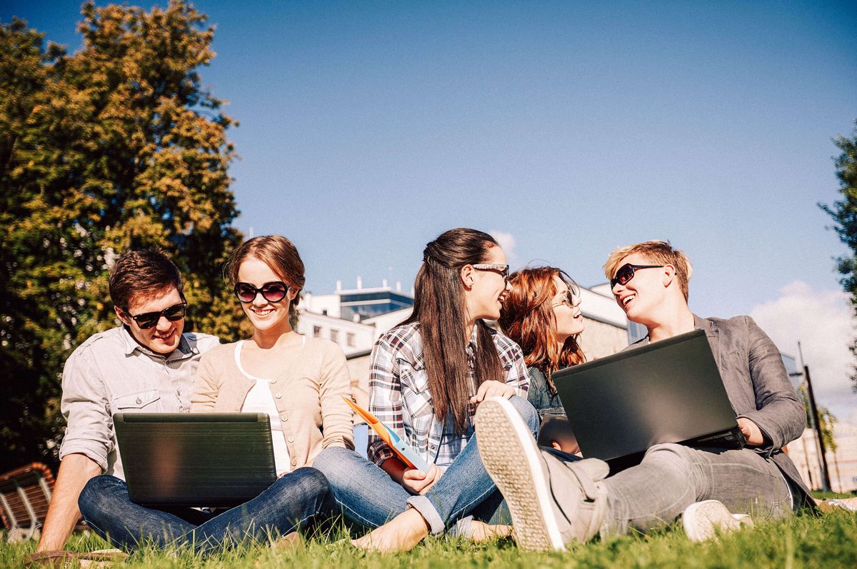 zur Vergrößerungsansicht des Bildes: Zwei junge Männer und drei junge Frauen mit Sonnenbrillen und zwei Laptops an einem sonnigen Tag auf einer Wiese sitzend und arbeitend.