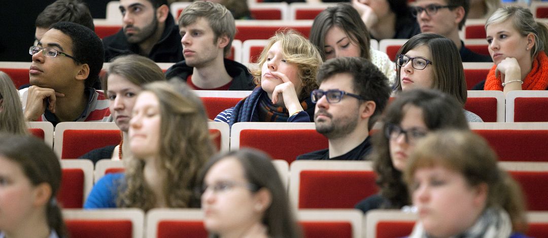 Mehrer Studierende sitzen im Hörsaal in roten Stuhlreihen