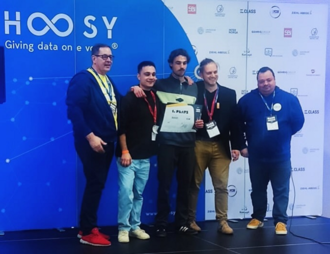 Das Gewinnerterteam (bestehend aus drei Personen) steht zwischen zwei Veranstaltern des Hoosy Hackathons und präsentiert die Urkunde des 1. Platzes