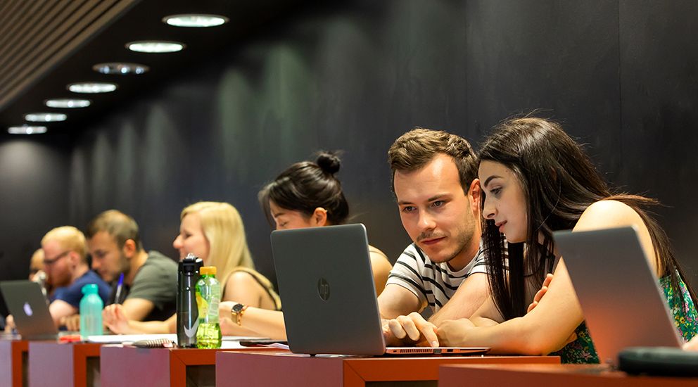 Studierende sitzen zusammen an den Arbeitsplätzen im Hörsaalgebäude und schauen in einen Laptop.