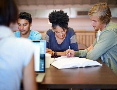 Studierende sitzen an einem Tisch in der Bibliothek und lernen gemeinsam. Auf dem Tisch liegen Bücher und Computer.