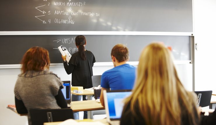 Lehrerin schreibt etwas an die Tafel, während Schüler und Schülerinnen aufmerksam schauen. Foto: Colourbox