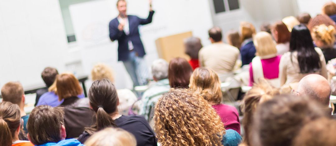 Im Vordergrund größeres Publikum von hinten, im Hintergrund ein Sprecher vor der Wand