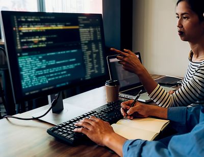 Farbfoto: Ein Mann und eine Frau sitzen an einem Computerarbeitsplatz und blicken auf einen Monitor, der Programmiercode anzeigt. Die Frau zeigt auf den Monitot. Der Mann tippt mit der linken Hand und macht sich mit der rechten Hand Notizen in einem Buch.