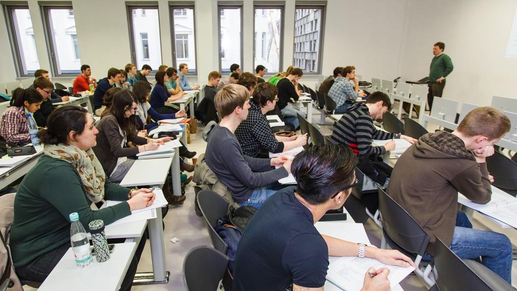 zur Vergrößerungsansicht des Bildes: Studierende in einem Seminarraum verfolgen die Vorlesung eines Dozenten im Institutsgebäude.