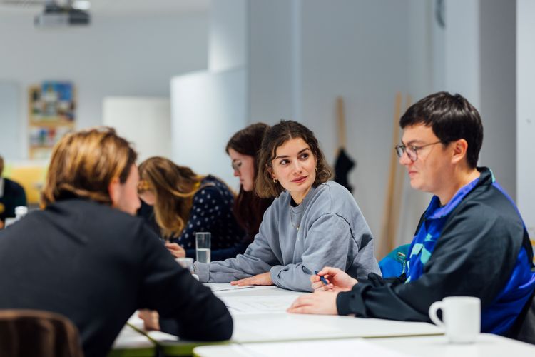 Farbige Aufnahme von drei Personen, die sich gegenüber an einem Tisch sitzen und miteinander sprechen; im Hintergrund sind weitere an Tischen sitzende Personen verschommen erkennbar