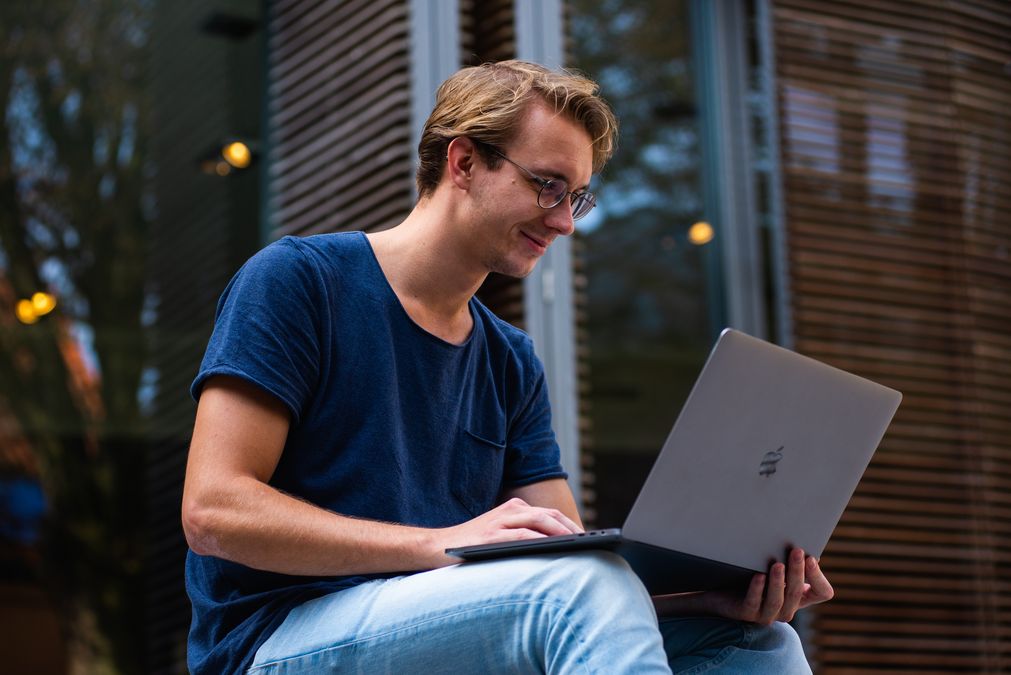 zur Vergrößerungsansicht des Bildes: Ein junger Mann mit Brille und einem Laptop auf dem Schoß vor einem Gebäude sitzend