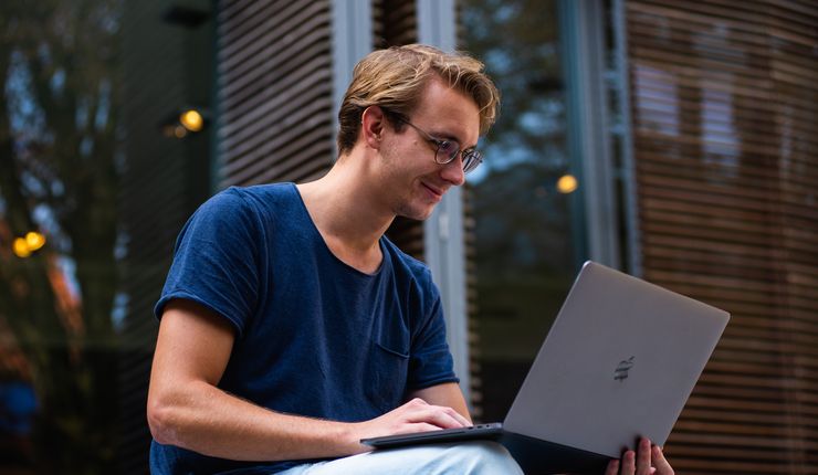 Junger Mann mit Laptop auf dem Schoß vor einem Gebäudeeingang