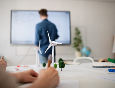 Im Vordergrund ist ein Schreibtisch, auf dem ein Modellwindrad steht. Im Hintergrund steht eine Lehrperson vor einer Tafel und unterrichtet die Personen am Tisch.