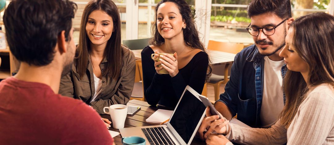 Five people at a coffee table with laptops, cups and paper are having a discussion.