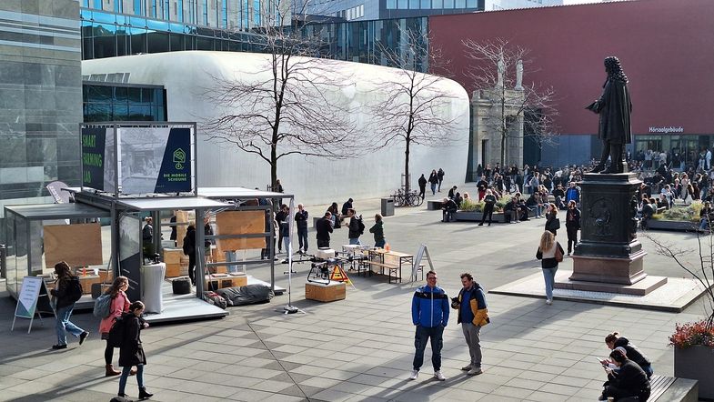 Farbfoto: Blick den Innenhof am Campus Augustusplatz, Messestand der mobilen Schule ist aufgebaut, einige Menschen sind im Gespräch mit einander und schauen sich die Angebote an