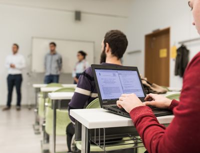 Studierende halten eine Vortrag vor der Gruppe, während die Person im Fokus des Bildes Notizen auf dem Laptop macht.