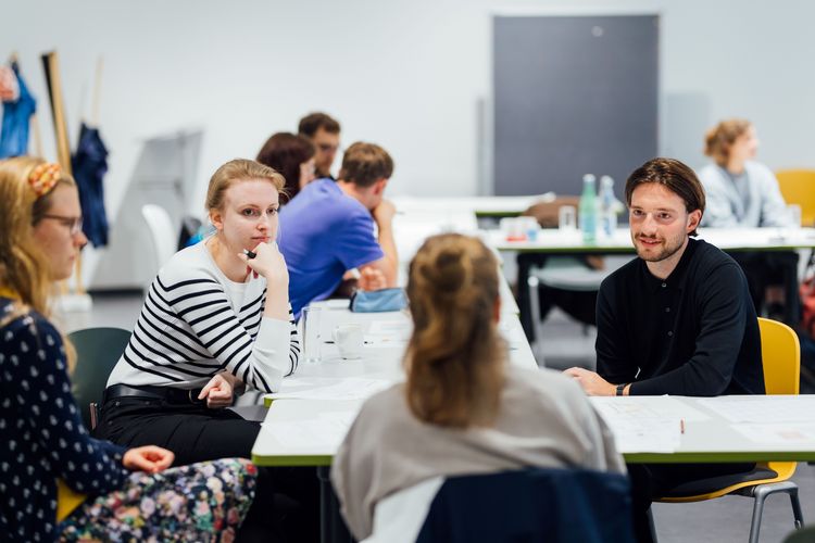 Farbige Aufnahme von vier Personen im Gespräch, die um einen Tisch herum sitzen; im Hintergrund sind weitere an Tischen sitzende Personen vreschwommen zu erkennen