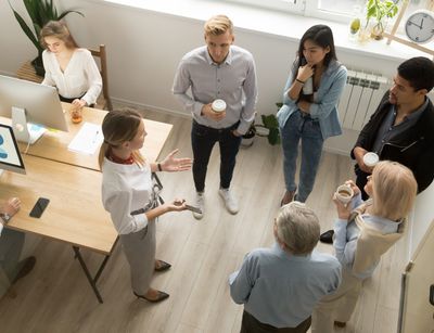 Six people stand in a circle for a casual meeting, a woman tells them something.