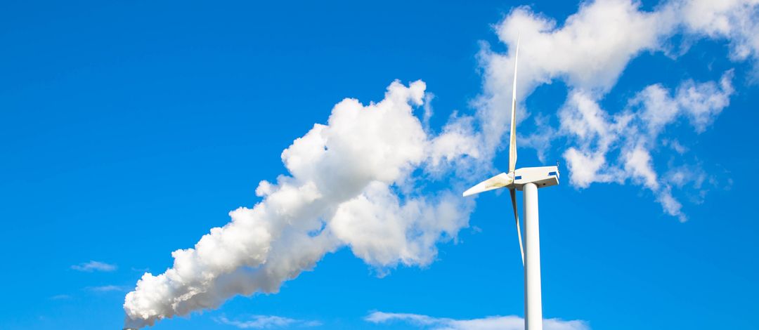 Rauchender Schlot und rotierende Windkraftanlage zueinander gewendet vor blauem Himmel.
