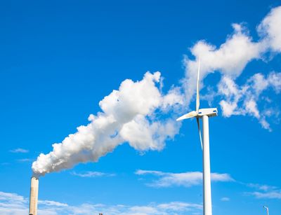 Rauchender Schlot und rotierende Windkraftanlage zueinander gewendet vor blauem Himmel.