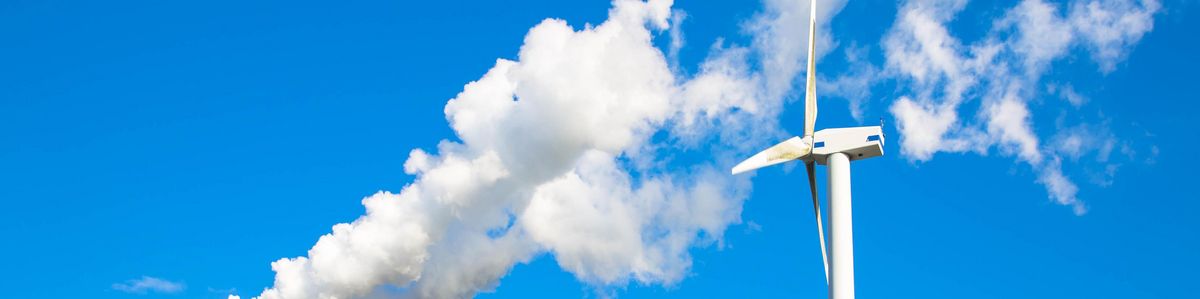 zur Vergrößerungsansicht des Bildes: Rauchender Schlot und rotierende Windkraftanlage zueinander gewendet vor blauem Himmel.