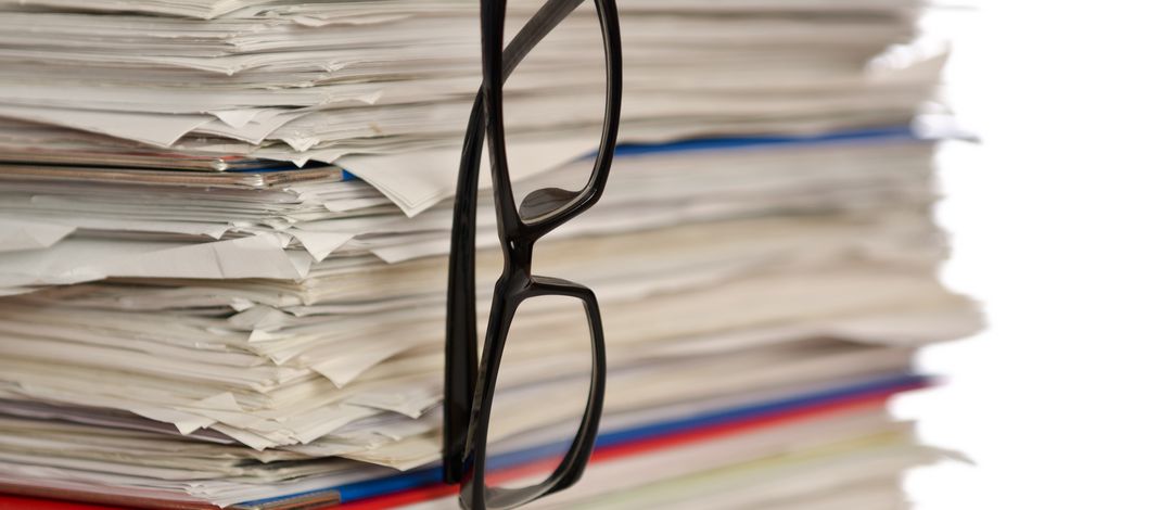 A stack of papers, with reading glasses hanging down.